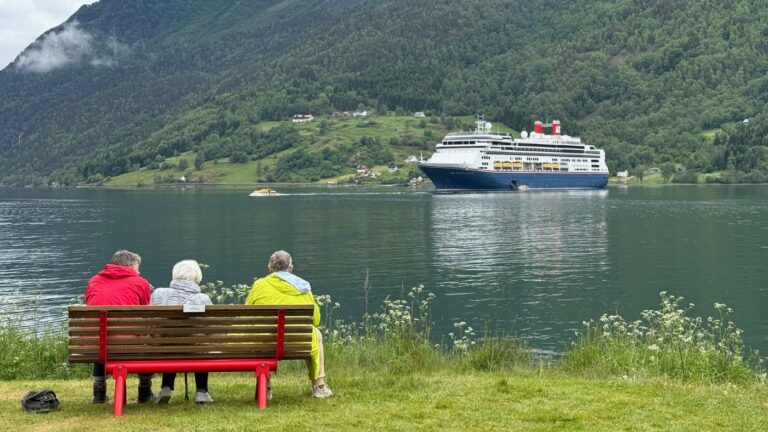 Trois personnes en manteaux clairs sur un banc à Skjolden regardant un bateau de croisière dans le fjord. Photo : David Nikel.